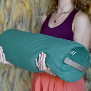 Kobieta w fioletowym topie i pomarańczowej spódnicy trzyma w ramionach duży zielony wałek do jogi WellB, tło stanowi ściana z roślinnym wzorem.